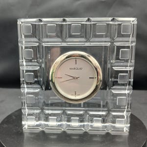 Puede incluir: Un reloj cuadrado de cristal transparente con un patrón de rejilla geométrica en el marco. La esfera circular es plateada con manecillas negras y el texto MARQUIS impreso en el centro. El reloj está incrustado en un bloque de cristal grueso.