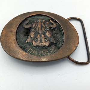 May include: A bronze belt buckle featuring a raised image of a bull's head with the word "TAURUS" inscribed below. The buckle is attached to a brown leather belt.