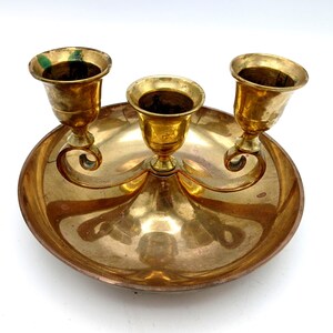May include: A vintage brass candle holder with three candle cups. The candle holder has a round base with a decorative design and curved arms supporting the candle cups. The brass has a warm, golden color.