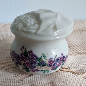 May include: A white ceramic jar with a floral design. The jar has a lid with a ruffled edge and a floral design. The jar is decorated with purple and white flowers.