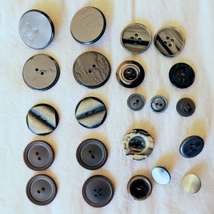 Puede incluir: Surtido de 18 botones en varios colores, patrones y materiales, incluyendo negro, marrón, blanco y gris. Algunos botones tienen una superficie texturizada, mientras que otros son lisos. Los botones están dispuestos en un patrón de cuadrícula sobre un fondo de tela blanca.