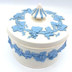 May include: A white ceramic round trinket box with a lid, decorated with blue grape vine relief detailing. The lid has a conical finial with blue vertical stripes. The box has a matching blue grape vine design around the base.