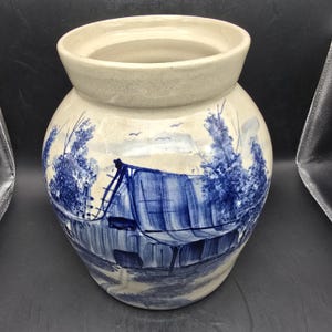 Puede incluir: Un jarrón de cerámica con un borde ancho y acampanado y un cuerpo redondeado. El jarrón está pintado con una escena de paisaje azul y blanco con un edificio y árboles. El borde es de color beige claro.
