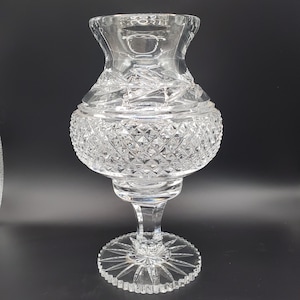 Puede incluir: Un jarrón de vidrio transparente con un diseño de cristal tallado. El jarrón tiene una base ancha y un cuello estrecho. Es perfecto para exhibir flores u otros artículos decorativos.