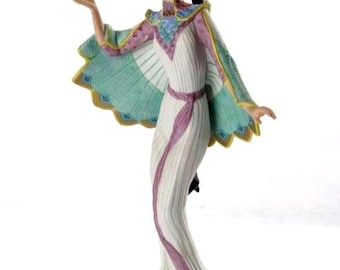 Puede incluir: Figura de porcelana de una mujer con un vestido fluido y una capa turquesa y amarilla. La figura tiene una corona y está en una pose elegante. El vestido es blanco con detalles morados.