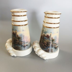 May include: Pair of white porcelain candle holders with a seascape design featuring a lighthouse and a pier. The holders have gold accents and a textured base.