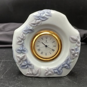 May include: White porcelain clock with a gold rim and a Lladró logo. The clock face has Roman numerals and a gold-colored hour and minute hand. The clock is decorated with blue and white floral designs.