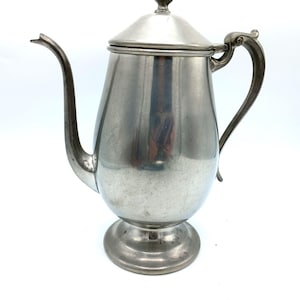 Puede incluir: Una cafetera vintage de metal plateado con un pico curvo y un asa decorativa. La cafetera tiene una tapa y se asienta sobre una base de pedestal. El diseño es clásico y elegante, adecuado para servir café o té.