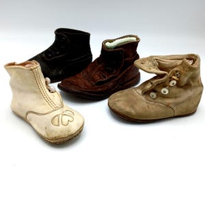 May include: Four antique baby shoes in various styles and colors. The shoes include a black leather boot, a brown leather boot, a tan suede shoe with button closures, and an off-white shoe with a decorative design.