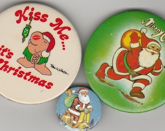 May include: Three round Christmas themed buttons. The first button is white with red text that says "Kiss Me... it's Christmas" and features a cartoon character with a green outfit and red hat. The second button is green with white text that says "Merry Christmas" and features a cartoon Santa Claus carrying a bag of gifts. The third button is blue with white snowflakes and features a cartoon Santa Claus riding a scooter.