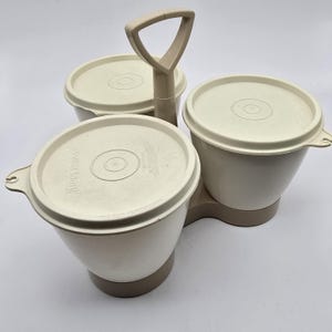 May include: A vintage Tupperware serving set in beige. The set includes three round bowls with lids, connected by a base and a handle. The lids and bowls are a light beige, with a darker beige base. The handle is triangular.