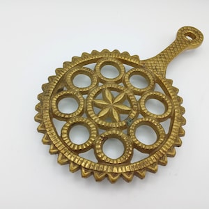 Puede incluir: Un salvamanteles de metal dorado con asa. El salvamanteles presenta un diseño circular con una estrella en el centro, rodeada de círculos y un borde decorativo. El asa tiene un patrón texturizado.