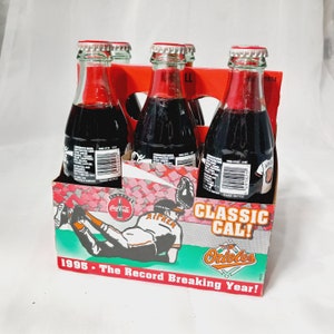 May include: Six-pack of Coca-Cola Classic soda bottles in a cardboard box with a Baltimore Orioles theme. The box features a cartoon image of Cal Ripken Jr. in a baseball uniform and the text "Classic Cal! Orioles 1995 - The Record Breaking Year!"