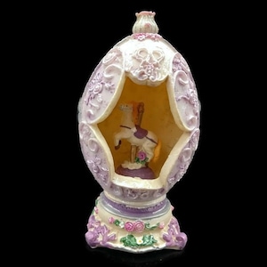May include: A white and purple ceramic egg-shaped music box with a carousel horse inside. The music box has a floral design and a base with a floral design.