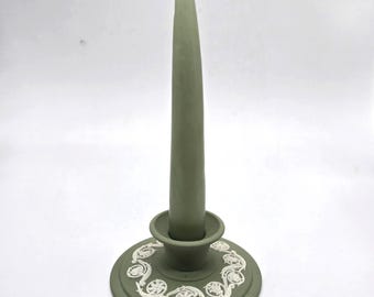 May include: A sage green candlestick with a matching taper candle. The candlestick has a round base with a white, ornate design. The candle is approximately 8 inches tall.