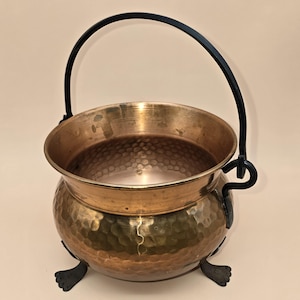 May include: A hammered copper cauldron with a black metal handle and feet. The cauldron has a round body and a wide rim. The handle is arched and attached to the cauldron with decorative hooks. The cauldron is likely used for decorative purposes.