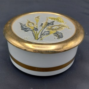 Könnte beinhalten: Eine weiße Keramik-Schmuckdose mit goldfarbenem Rand und einem goldfarbenen Band um die Mitte. Der Deckel zeigt ein silbernes und goldenes Reliefdesign mit Blumen und einem Schmetterling.