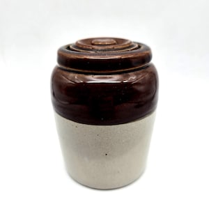 Könnte beinhalten: Ein Vintage-Steinzeugtopf mit Deckel. Der Topf hat ein zweifarbiges Design, mit einem dunkelbraunen oberen Bereich und einem hellbeigen unteren Bereich. Der Deckel ist ebenfalls dunkelbraun. Dieses antike Gefäß wird wahrscheinlich zur Aufbewahrung von Lebensmitteln verwendet.