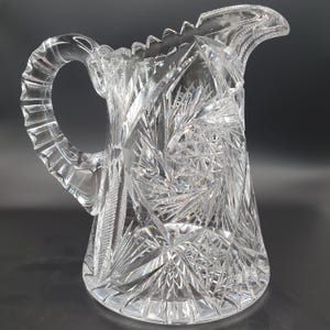 Puede incluir: Una jarra de cristal tallado transparente con un asa grande y un pico. La jarra presenta intrincados patrones de estrellas y geométricos en toda su superficie. El borde tiene un diseño de dientes de sierra. La jarra probablemente se utiliza para servir bebidas.