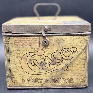 May include: Vintage yellow metal Cinco Handy Humidor with a hinged lid, handle, and clasp. The box features the word "Cinco" in a decorative font and text that reads "The cigars herein contained were manufactured to retail at more than five cents each and not more than eight cents each and are so tax-paid."