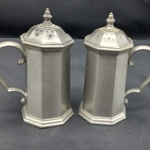 May include: A set of two silver pewter salt and pepper shakers with octagonal bodies and handles. The shakers have a decorative design on the top and a small hole for dispensing salt and pepper.