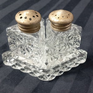 May include: A set of two clear glass salt and pepper shakers with silver tops, sitting on a matching clear glass base with a diamond pattern.