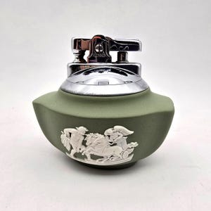 May include: An olive green Ronson lighter with a silver-colored top. The lighter features a white bas-relief design of figures and a chariot. The lighter is a vintage collectible.