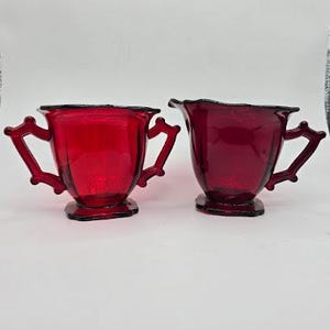 May include: A set of two vintage red glass creamer and sugar bowl with ornate handles. The creamer has a pouring spout and the sugar bowl has a lid.