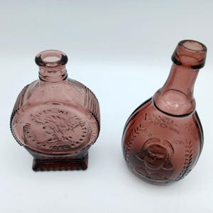 May include: Two vintage glass bottles with embossed designs. One bottle is purple and features the words "In God We Trust" and "1959". The other bottle is a darker purple and features the words "Johnny Fedora" and a portrait of a man.