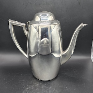 May include: A silver-colored coffee pot with a curved spout and a handle. The pot has a lid and a rounded body. The design is Art Deco style, with a polished finish. The pot is made of metal and is suitable for serving coffee.