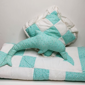 Puede incluir: Una colcha de retazos turquesa y blanca con un juguete de peluche de delfín turquesa en la parte superior. La colcha está hecha de cuadrados con un patrón floral.