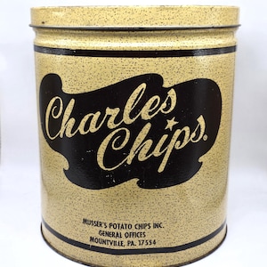 May include: Vintage Charles Chips tin with a speckled tan background and black accents. The tin features the words "Charles Chips" in a stylized font within a black oval. The bottom of the tin includes text from Musser's Potato Chips Inc.