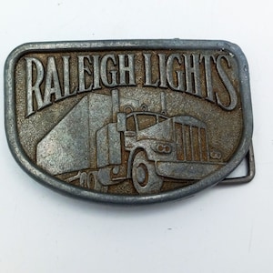 May include: A silver metal belt buckle with the words "Raleigh Lights" and a depiction of a semi-truck.