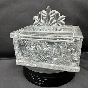 May include: A clear glass rectangular box with a lid featuring a prominent, upright, fan-shaped crystal finial. The exterior surfaces of the box and lid are decorated with intricate, pressed geometric starburst and snowflake patterns.
