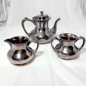 May include: A silver tea set with a teapot, creamer, and sugar bowl. Each piece has a beaded rim and a monogrammed design.
