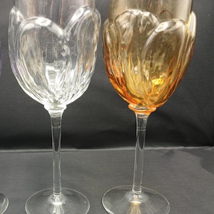 May include: Two stemmed wine glasses with a textured, petal-like exterior design. One glass is made of clear, transparent glass, while the other is crafted from amber-colored glass. Both feature a long, slender stem and a wide, circular base.