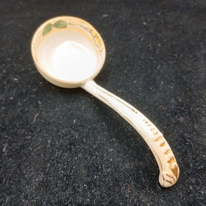 May include: A white ceramic ladle with a rounded bowl and a curved handle. The rim of the bowl features a floral design in yellow and green. Gold accents adorn the handle. The ladle is designed for serving.