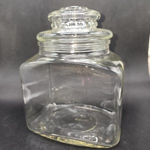 May include: A clear glass jar with a square base and a rounded lid. The jar has a wide opening and a decorative, textured lid. The jar is empty and sits against a dark background. The jar is likely used for storage.