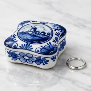 May include: A small, square, white ceramic trinket box featuring blue hand-painted floral patterns and a central circular illustration of a windmill. A simple, polished silver-toned metal band ring is positioned next to the ceramic box.