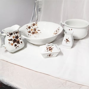 May include: A white porcelain wash set featuring a pitcher, bowl, soap dish, small vase, and chamber pot. Each piece is decorated with a brown and tan floral pattern. The set is displayed on a white surface.