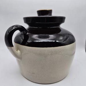 May include: A vintage stoneware jug with a black top and handle, and a cream-colored base. The jug has a lid and a curved handle. The ceramic piece is likely used for storing liquids or as a decorative item.