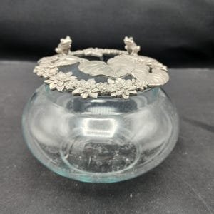 May include: A clear glass bowl with a decorative metal lid. The lid features a ring of flowers, lily pads, and two small frog figurines. The bowl is round and has a slight blue tint.