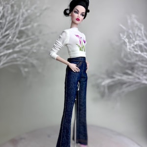 Op de afbeelding: Een modepop met een witte top met lange mouwen met een paars bloemenmotief en een donkerblauwe flared jeans. De pop heeft zwart haar in een knot en staat op een transparante standaard.