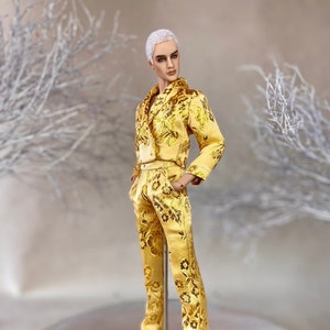 May include: A doll in a gold suit with a floral design. The outfit comprises a double-breasted jacket and trousers. The doll is also wearing black shoes. The doll has short white hair and is standing on a clear stand. The suit's measurements are in centimetres.