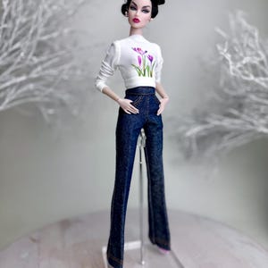 Op de afbeelding: Een modepop met een witte top met lange mouwen met een paars bloemenmotief en een donkerblauwe flared jeansbroek. De pop heeft zwart haar en staat op een transparante standaard. De pop draagt roze schoenen.