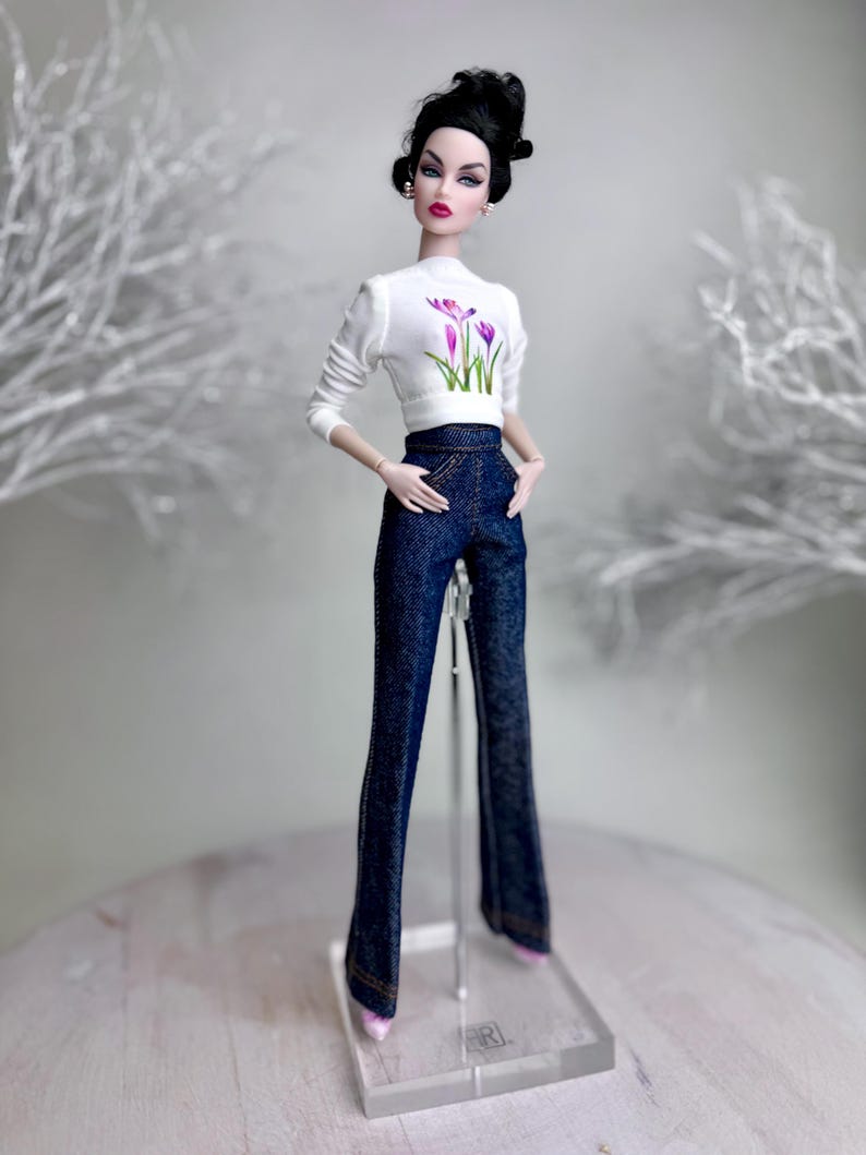Op de afbeelding: Een modepop met een witte top met lange mouwen met een paars bloemenmotief en een donkerblauwe flared jeansbroek. De pop heeft zwart haar en staat op een transparante standaard. De pop draagt roze schoenen.
