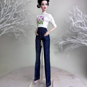 Op de afbeelding: Een modepop draagt een witte top met lange mouwen met een bloemenontwerp en een donkerblauwe spijkerbroek. De pop heeft zwart haar en staat op een transparante standaard. De pop draagt roze schoenen en oorbellen.