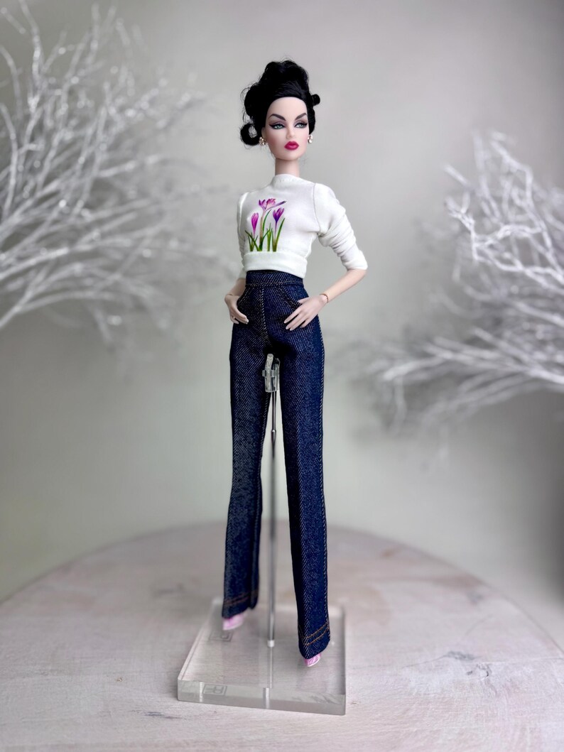 Op de afbeelding: Een modepop draagt een witte top met lange mouwen met een bloemenontwerp en een donkerblauwe spijkerbroek. De pop heeft zwart haar en staat op een transparante standaard. De pop draagt roze schoenen en oorbellen.