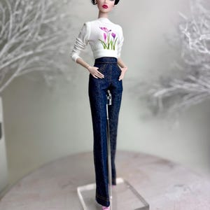 Op de afbeelding: Een modepop met een witte top met lange mouwen met een bloemenontwerp en een donkerblauwe spijkerbroek. De pop heeft donker haar en draagt oorbellen en roze schoenen. Ze staat op een transparante standaard. De outfit is stijlvol.