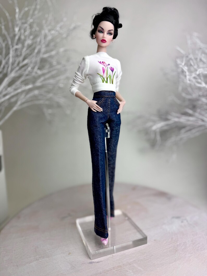 Op de afbeelding: Een modepop met een witte top met lange mouwen met een bloemenontwerp en een donkerblauwe spijkerbroek. De pop heeft donker haar en draagt oorbellen en roze schoenen. Ze staat op een transparante standaard. De outfit is stijlvol.